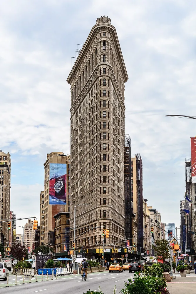 The narrow wedge-shaped Flatiron Building on a cloudy day, with a cyclist and taxis at street level